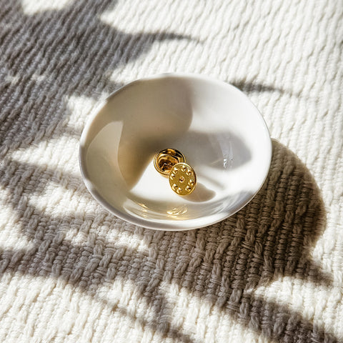 Golden butterfly clasp in a small white dish on a textured cream rug, sunlight casting soft shadows.