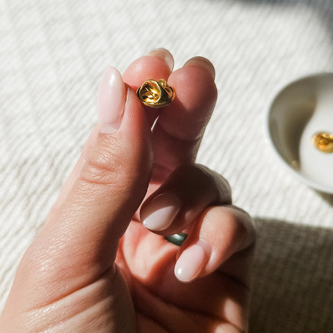 Close-up of a hand holding a golden butterfly clasp, with a soft background of a white dish and textured cream rug.