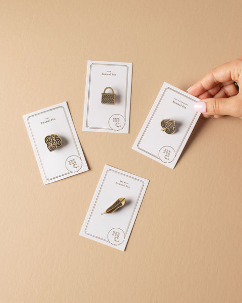 Gold Māori pins on cards with a hand holding one of the pins against a beige background