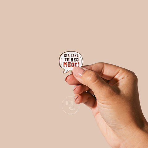  A hand holding an enamel pin with the words "Kia Kaha Te Reo Māori" written in bold letters against a neutral-toned background.