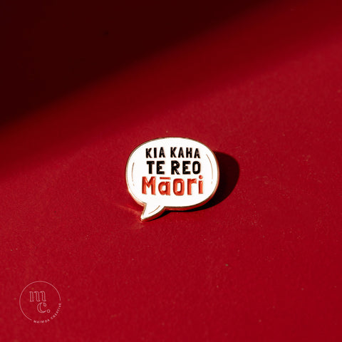  An enamel pin with the inscription "Kia Kaha Te Reo Māori" placed on a bold red background, with clear white, red, and black colours and brass detailing around the edges.