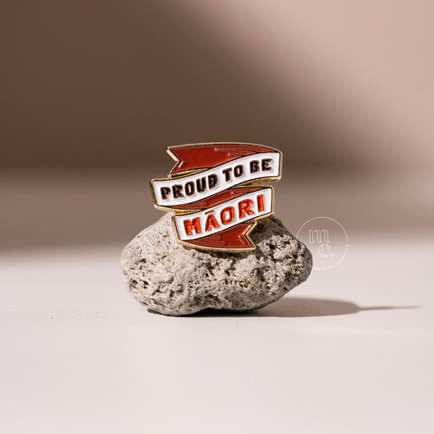  An enamel pin in a banner shape with the inscription "Proud to be Māori" placed on a textured pumice stone, with clear white, red, and black colours and brass detailing around the edges.