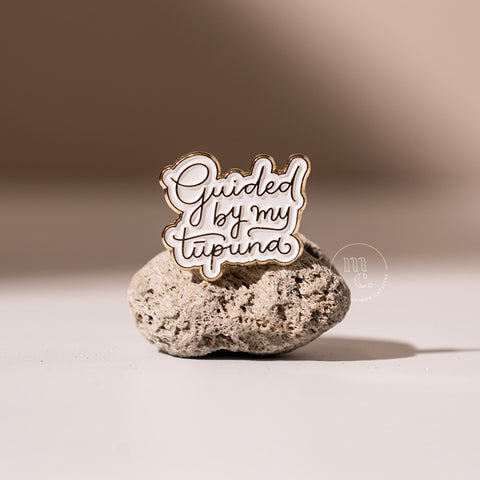  An enamel pin with the inscription "Guided by my Tūpuna" placed on a textured pumice stone, with clear white and brass colours and brass detailing around the edges.
