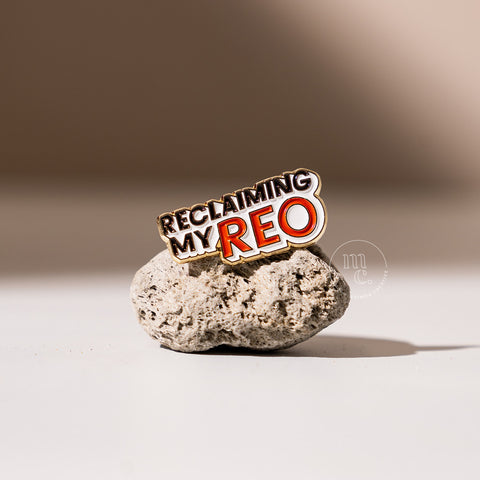  An enamel pin with the inscription "Reclaiming my Reo" placed on a textured pumice stone, with clear white, red, and black colours and brass detailing around the edges.