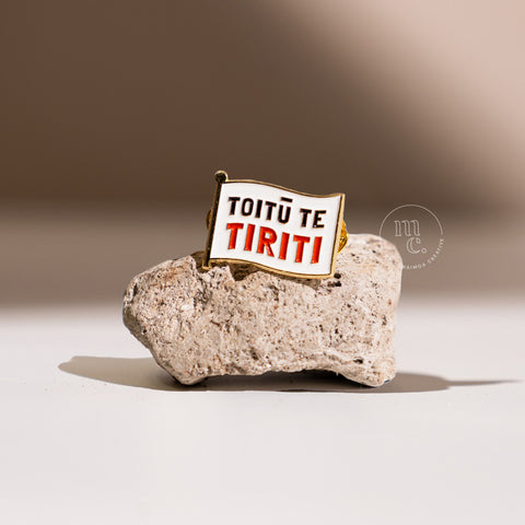  An enamel pin with the inscription "Toitū Te Tiriti" placed on a textured pumice stone, with clear white, red, and black colours and brass detailing around the edges.