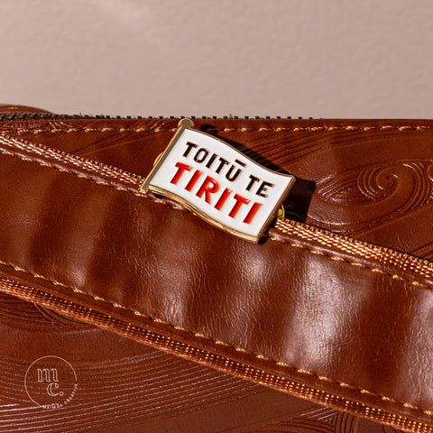  An enamel badge pin with the words "Toitū Te Tiriti" written in bold letters on a brown leather bag with Māori motifs and designs