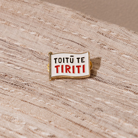  An enamel badge pin with the words "Toitū te Tiriti" written in bold letters on a natural textured material