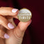 Hand holding a gold and white badge with 'TANGATA TIRITI' text against a blurred background