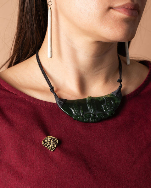 Close-up of a person wearing a pounamu and decorative tiki gold pin and a maroon top.