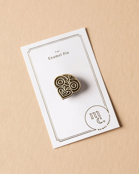 Gold tiki enamel pin on a white card with Maimoa Creative gold foil branding on a beige background