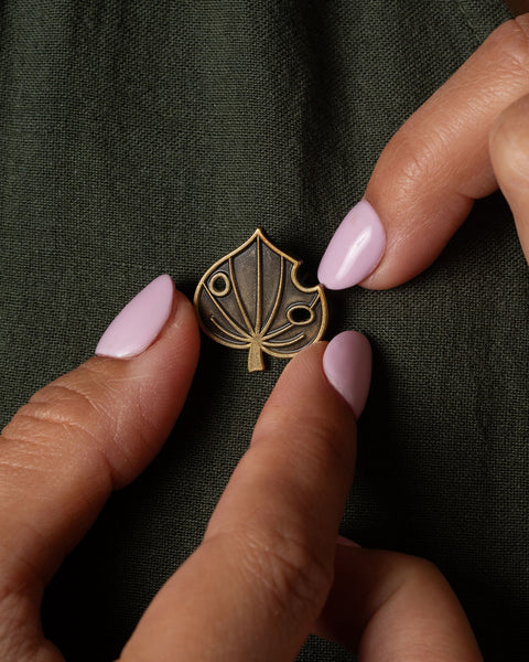 Gold kawakawa leaf brooch being held by a hand with pastel pink nail polish on a dark green fabric background