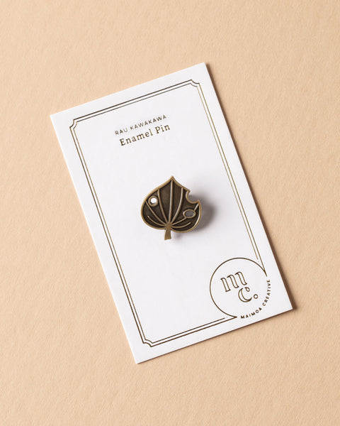 Gold kawakawa leaf enamel pin on a white card with Maimoa Creative gold foil branding on a beige background