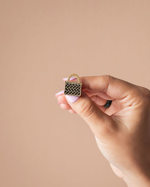 Hand holding a small gold kete basket pin against a beige background