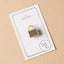 Gold kete enamel pin on a white card with Maimoa Creative gold foil branding on a beige background