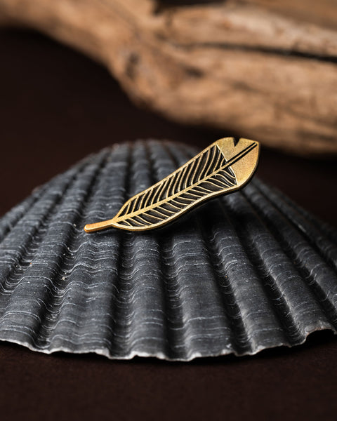 Gold huia feather brooch on a dark textured shell with a brown and wooden background