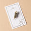 Gold huia feather enamel pin on a white card with Maimoa Creative gold foil branding on a beige background