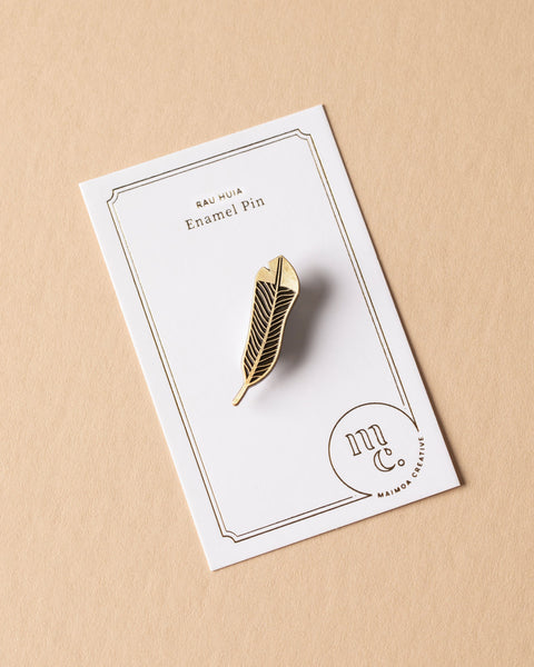 Gold huia feather enamel pin on a white card with Maimoa Creative gold foil branding on a beige background