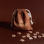 Fabric drawstring bag labelled 'Pakiaka' surrounded by scattered wooden Māori letter tiles on a deep brown background.