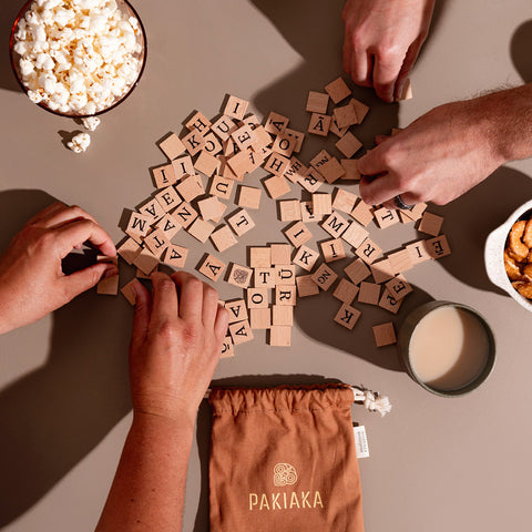 Hands assembling wooden letter tiles from the Pakiaka game on a table, accompanied by snacks and tea, with a drawstring bag on the side.