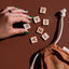 Hand with pounamu green nails arranging wooden Pakiaka game letter tiles next to a drawstring bag