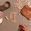 Hand assembling wooden letter tiles into a crossword from the Pakiaka game, accompanied by snacks and tea, with a drawstring bag on the side.