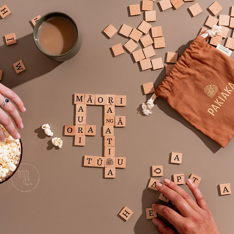 Hand assembling wooden letter tiles into a crossword from the Pakiaka game, accompanied by snacks and tea, with a drawstring bag on the side.