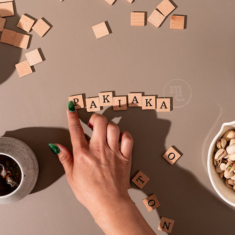 Hand assembling wooden letter tiles into the word 'Pakiaka', accompanied by a herb tea and pistachios, surrounded by more tiles.