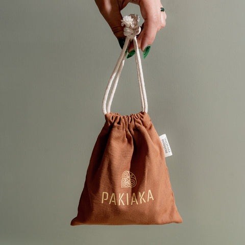 Manicured hand holding up a burnt orange cotton drawstring bag with the logo 'Pakiaka' on it with a earthy pastel green background