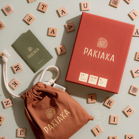 Flat lay of Pakiaka game with its box, instruction booklet, and tiles scattered on a light green background.