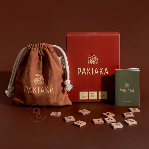 Pakiaka game set featuring a box, drawstring bag, instruction guide, and scattered letter tiles on a brown background.