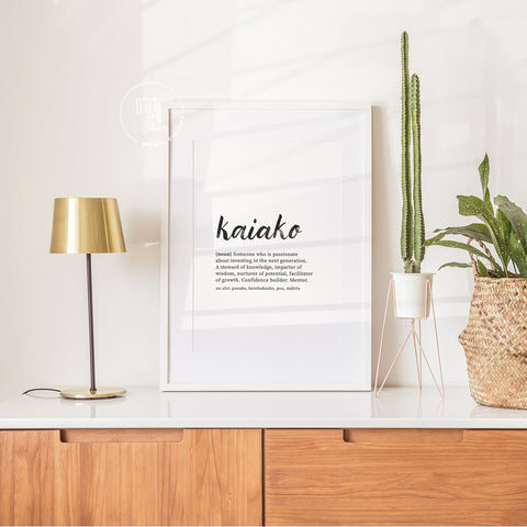 Framed Kaiako definition art print on white cabinet with modern lamp and indoor plants in a bright, sunlit room.