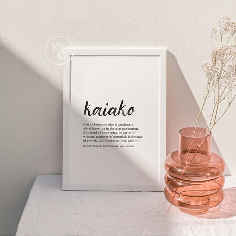 Framed A4 Māori definition art print of "Kaiako" displayed in soft natural light beside a glass vase and dried flowers.