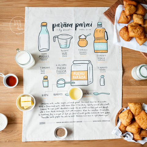 A spread of ingredients for making Māori fry bread, displayed around an illustrated recipe on a tea towel, with golden syrup, milk, butter, yeast, sugar, oil, and a plate of freshly made fry bread, all laid out on a wooden surface.