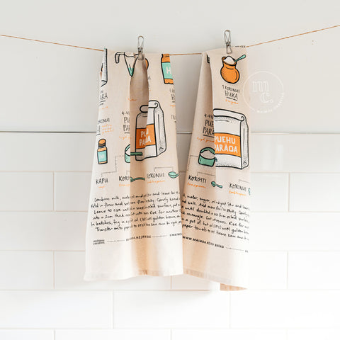 Two tea towels with illustrations and text from a Māori fry bread recipe are hanging on hooks against a white tiled kitchen wall, creating a cozy, culinary-themed decor.