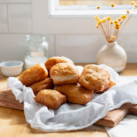 A pile of golden-brown Māori fry bread on a wooden cutting board, wrapped in a white cloth, with a glass jar, a bowl, and a vase with dried flowers in the background, on a kitchen counter by a window.