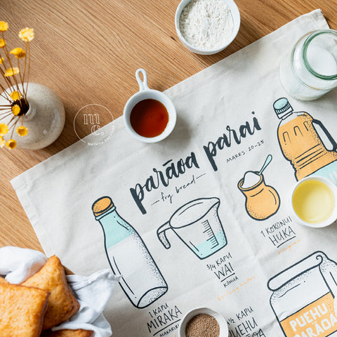 Overhead view of a tea towel with an illustrated recipe for Māori fry bread, accompanied by ingredients including flour, sugar in a jar, oil in a small dish, and a ramekin of golden syrup, with a vase of yellow flowers in the background, all on a wooden surface.