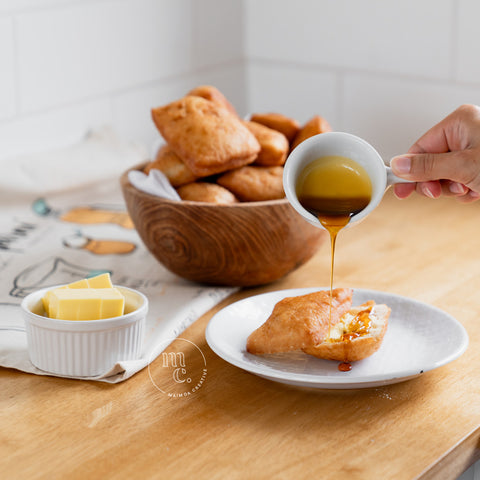 A close-up of a hand pouring golden syrup from a small white pitcher onto a piece of Maori fry bread on a white plate, with a bowl of butter and more fry bread in a wooden bowl in the background, on a wooden kitchen countertop.