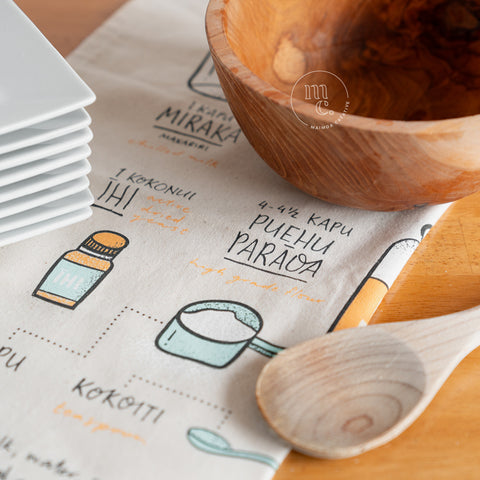 A tea towel with illustrated instructions for making Māori fry bread, partially covered by a stack of white plates, a wooden bowl, and a wooden spoon, arranged on a wooden kitchen surface.