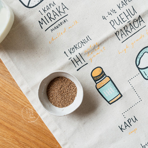 A tea towel with illustrated instructions for making Māori fry bread, partially covered by a small white bowl of yeast, on a wooden kitchen surface.