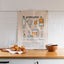 Kitchen scene with a tea towel featuring an illustrated Māori fry bread recipe hung on the wall, a wooden bowl, and a plate of fry bread on a wooden board on the countertop, with a subway tile backsplash in the background.