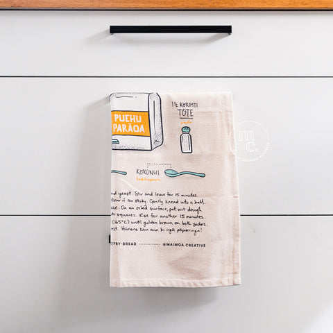 An illustrated tea towel with a recipe for fry bread hanging from a wooden drawer handle, featuring hand-drawn images of ingredients and instructions, against a white kitchen drawers with modern black handles.