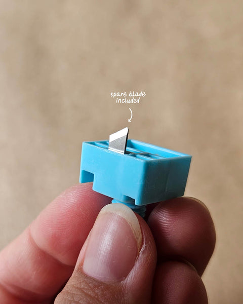 Blue blade holder for circle cutter, held between fingers against a beige background.