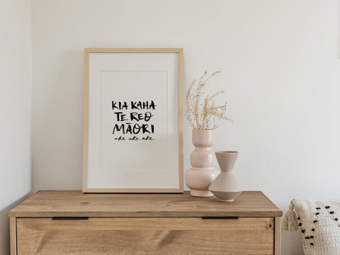 Framed print with hand-lettered text "Kia kaha te reo Māori ake ake ake" on a wooden shelf with decorative vases.