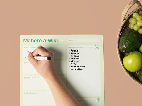 Hand writing on a magnetic A3 weekly planner with a black whiteboard marker, surrounded by green fruits on a beige background