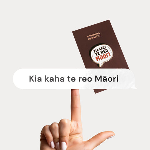 Hand holding a vinyl decal with "Kia Kaha Te Reo Māori" in a speech bubble design, with a bold enamel pin in the background with same message.