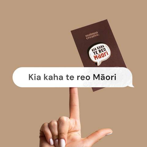 Hand holding a vinyl decal with "Kia Kaha Te Reo Māori" in a speech bubble design, with a bold enamel pin in the background with same message.