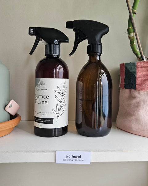 Two amber glass spray bottles on a white shelf, with the label 'kū horoi - cleaning products' in te reo māori stuck to the shelf