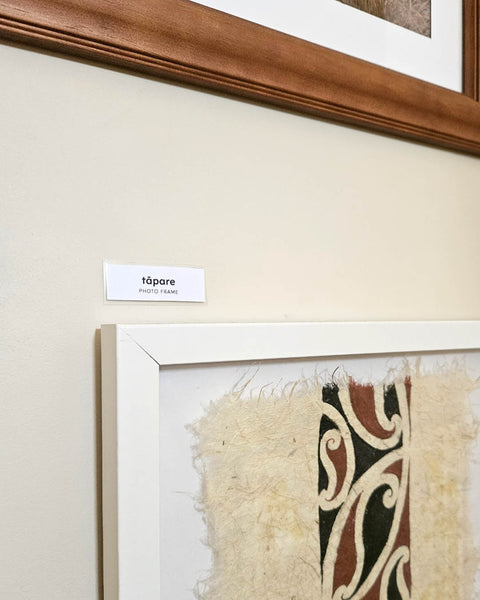 A framed Māori art piece with a white frame and a small label reading 'tāpare' in te reo Māori mounted on a wall