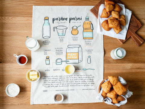 A spread of ingredients for making Māori fry bread, displayed around an illustrated recipe on a tea towel, with a cup of tea, milk, butter, yeast, sugar, oil, and a plate of freshly made fry bread, all laid out on a wooden surface.