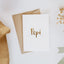 Flat lay of a premium reo Māori greeting card with the word 'Pēpi' in gold foil lettering, paired with a gold envelope and delicate dried flowers and baby muslin wrap with Māori motifs